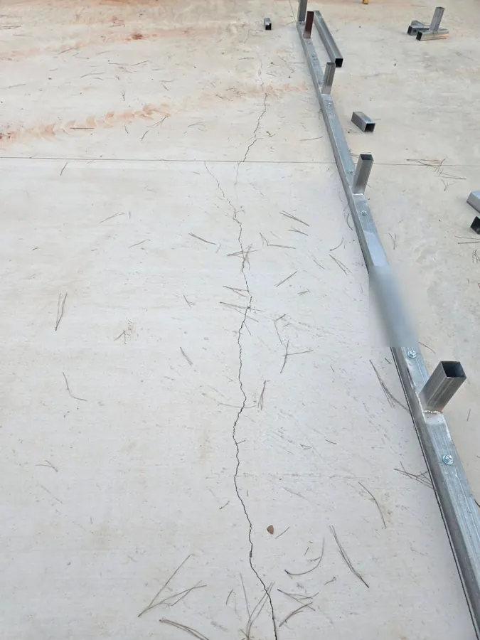 A long crack running across the concrete slab and through a saw-cut control joint, next to the steel base rail of the building